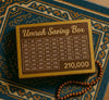 Wooden Umrah saving box with 210,000 goal, placed on patterned prayer mat with prayer beads